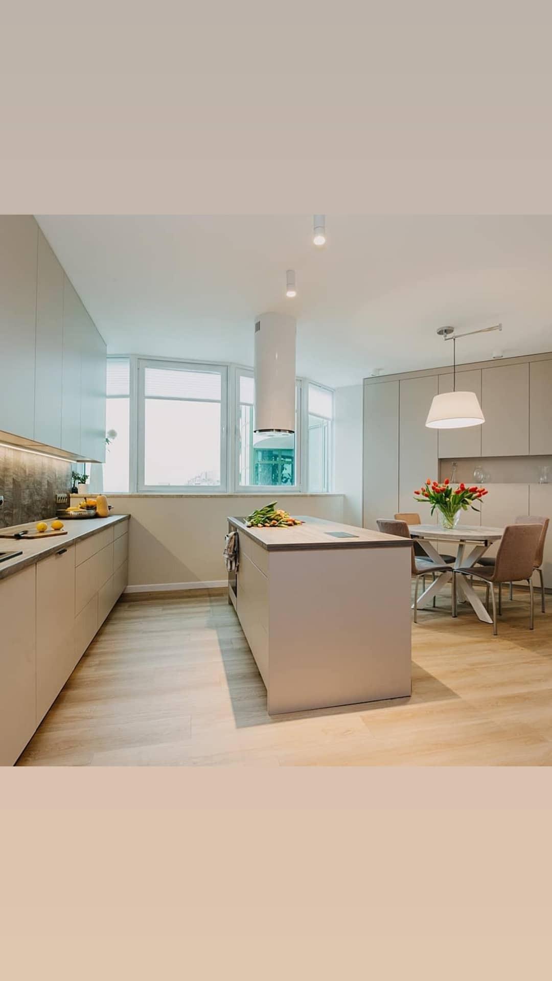 Jasna, przestronna kuchnia z wyspą, okrągłym stołem i bukietem czerwonych tulipanów. Minimalistyczne szafki, drewniana podłoga i nowoczesny okap cylindryczny.