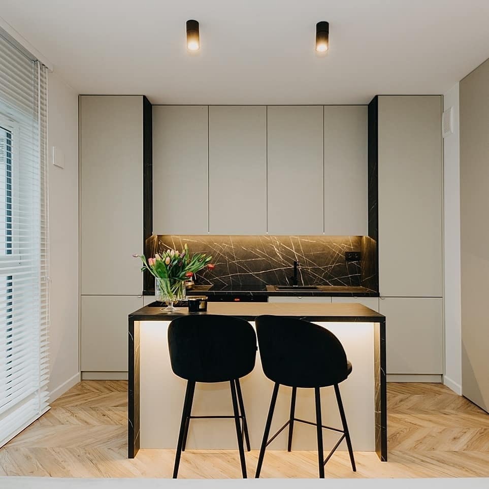 Nowoczesna kuchnia z wyspą, szare szafki, czarny blat z marmurowym wzorem, podświetlenie LED, dwie czarne, welurowe hokery, parkiet w jodełkę, bukiet tulipanów w szklanym wazonie.