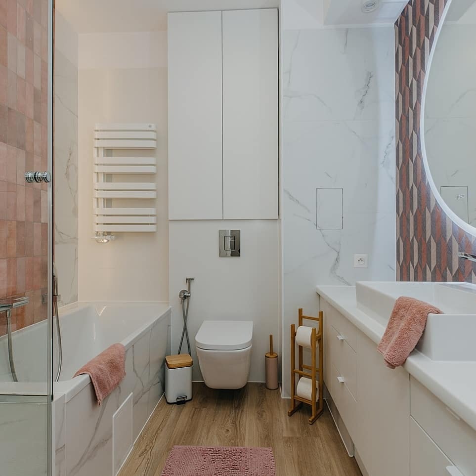 Nowoczesna łazienka z drewnianą podłogą, białą armaturą i akcentami w odcieniach różu, w tym ręczniki i dywanik. Na ścianie marmurowe płytki w połączeniu z geometrycznym wzorem przy okrągłym...