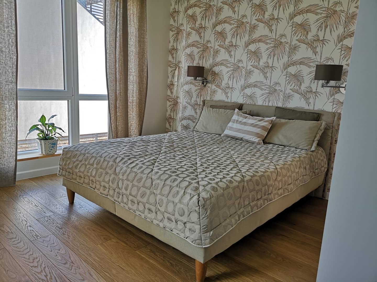 Sypialnia w beżach z łóżkiem z pikowaną narzutą, wzorzystą tapetą w palmy, drewnianą podłogą i kwiatem w doniczce na parapecie okna.