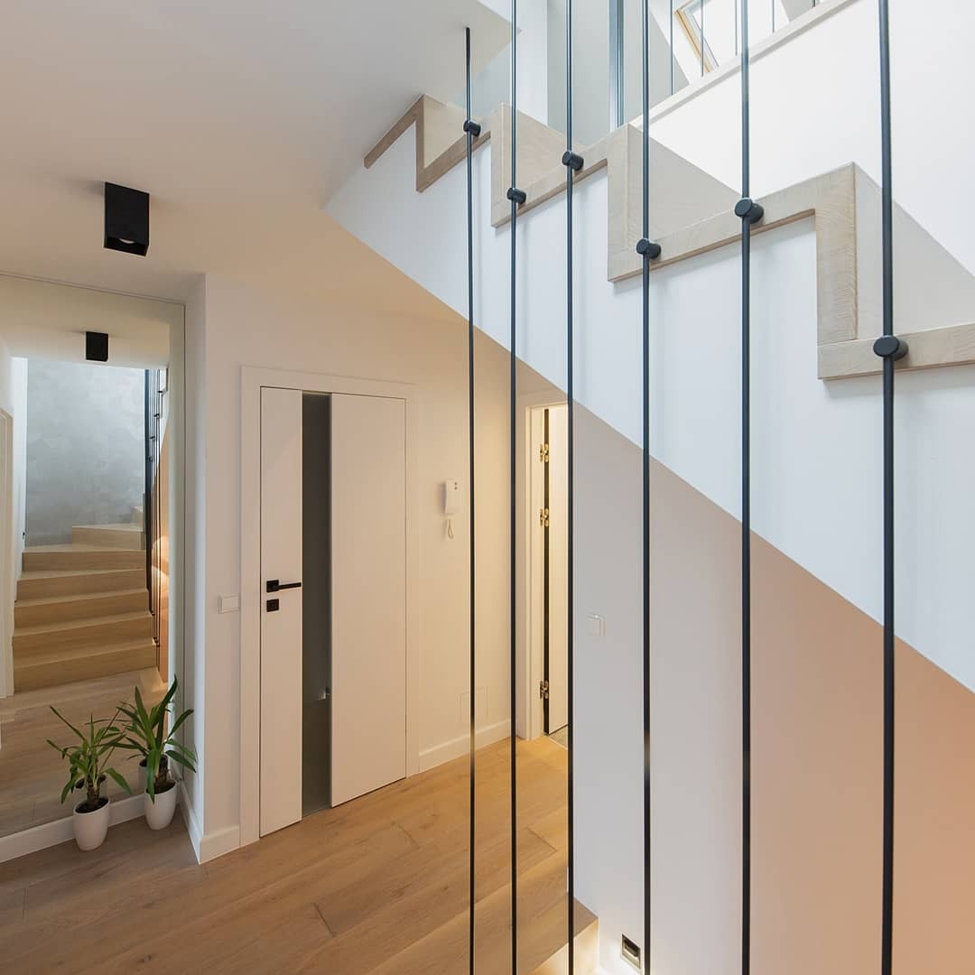 Nowoczesne wnętrze domu: jasne drewniane schody z czarnymi balustradami, białe ściany, drzwi z przeszkleniem i drewniana podłoga, widok w lustrze na kolejne schody.