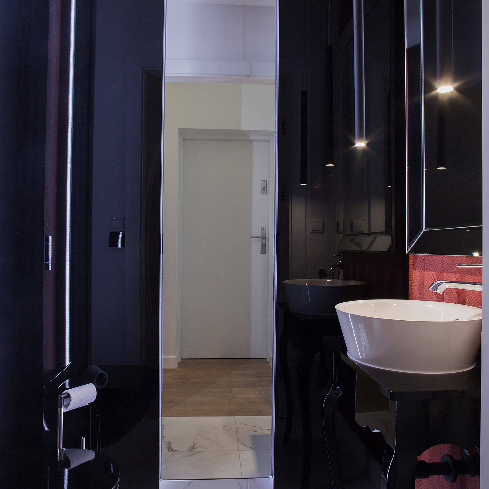 Nowoczesna łazienka z czarnymi, lustrzanymi ścianami, podwieszanymi lampami i umywalkami na dekoracyjnych, czarnych konsolach. W tle widoczna biała ściana i drzwi.