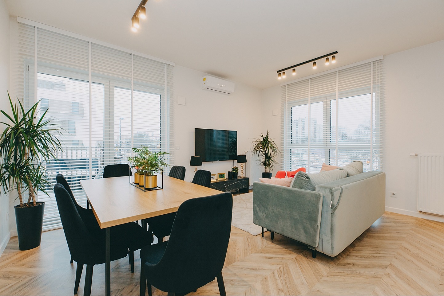 Jasne, przestronne wnętrze z drewnianą podłogą w jodełkę, dużym stołem z czarnymi krzesłami, zieloną sofą i roślinami doniczkowymi, oświetlone nowoczesnymi lampami sufitowymi i wpadającym światłem...