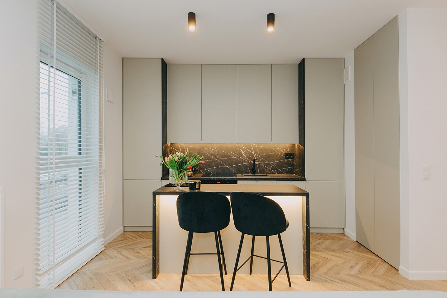 Nowoczesna kuchnia z wyspą, czarnymi hokerami i marmurowym blatem, oświetlona listwą LED pod szafkami wiszącymi, parkiet ułożony w jodełkę.