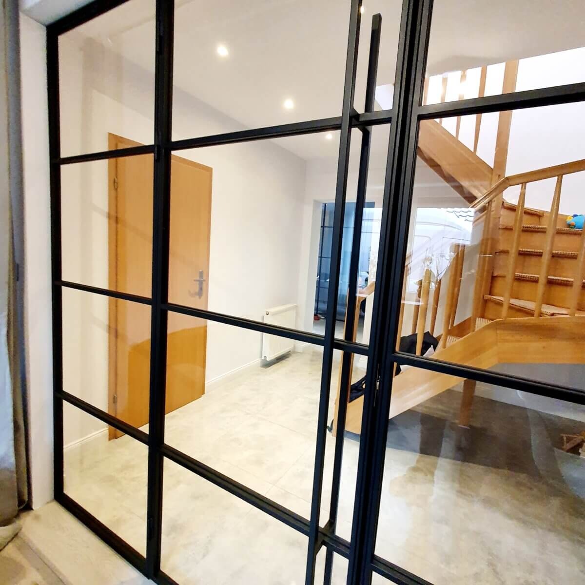 Szklana ścianka działowa z czarnymi profilami, widok na drewniane schody i drzwi w tle.