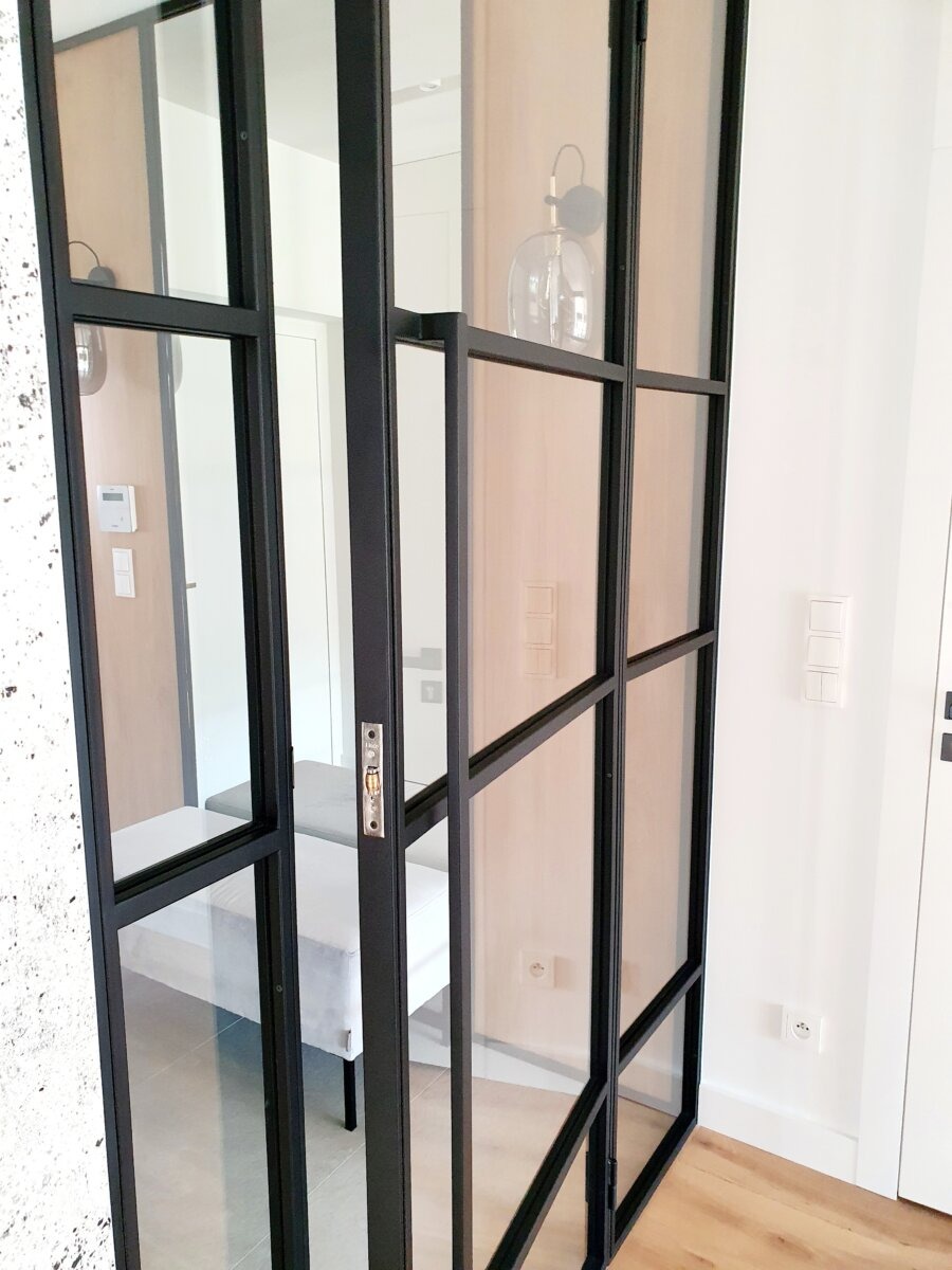 Nowoczesna, przeszklona konstrukcja z czarnymi profilami, dzieląca przestrzeń, widoczny fragment sypialni z szarą ławką, oświetlenie ścienne w formie szklanej kuli.