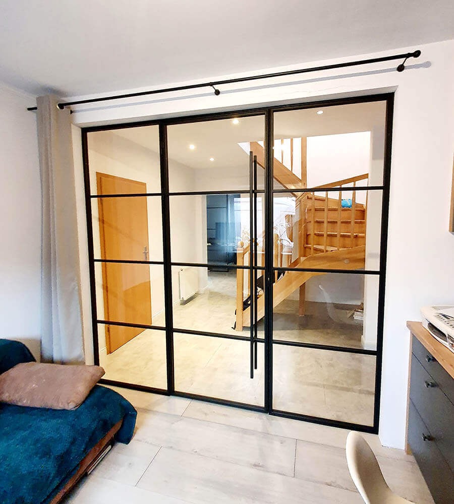 Szklane przegrody z czarnymi szprosami oddzielające przestrzeń, widok na drewniane schody i korytarz, nowoczesny design.