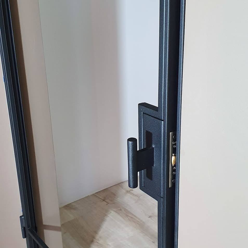 Czarne, nowoczesne drzwi z widoczną klamką i zamkiem, jasne ściany i podłoga z paneli w tle.