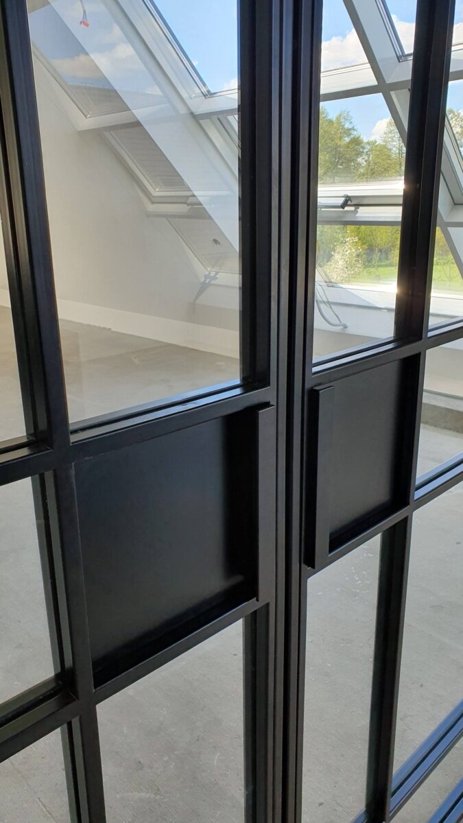 Czarne, metalowe, przeszklone skrzydła z widocznymi uchwytami na tle jasnego wnętrza z oknami dachowymi i widokiem na zieleń.