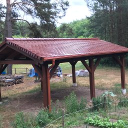 Solid Bud Damian Popławski - Drewniana altana z nowym, czerwono-brązowym dachem z dachówki ceramicznej, widoczna w otoczeniu drzew, z zapasem materiałów budowlanych w tle i zaparkowanym samochodem z otwartymi drzwiami.