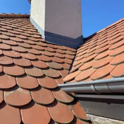 Dachówka karpiówka w odcieniach czerwieni, z widocznym kominem i szarą rynną, na tle błękitnego nieba.