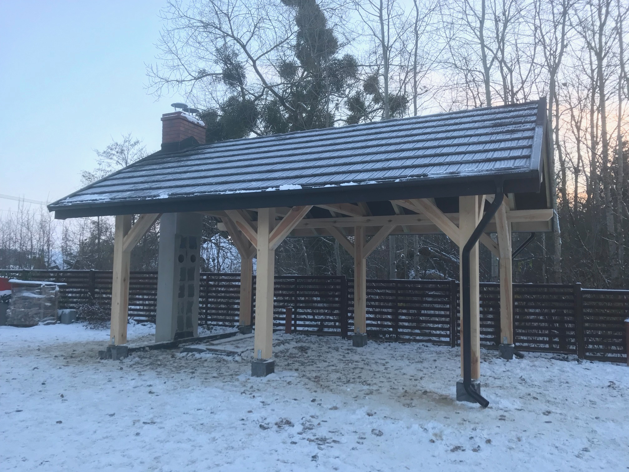 Drewniana konstrukcja z dachem z dachówki bitumicznej, częściowo pokryta śniegiem, z rynną i kominem z cegły, na zasypanym śniegiem terenie.