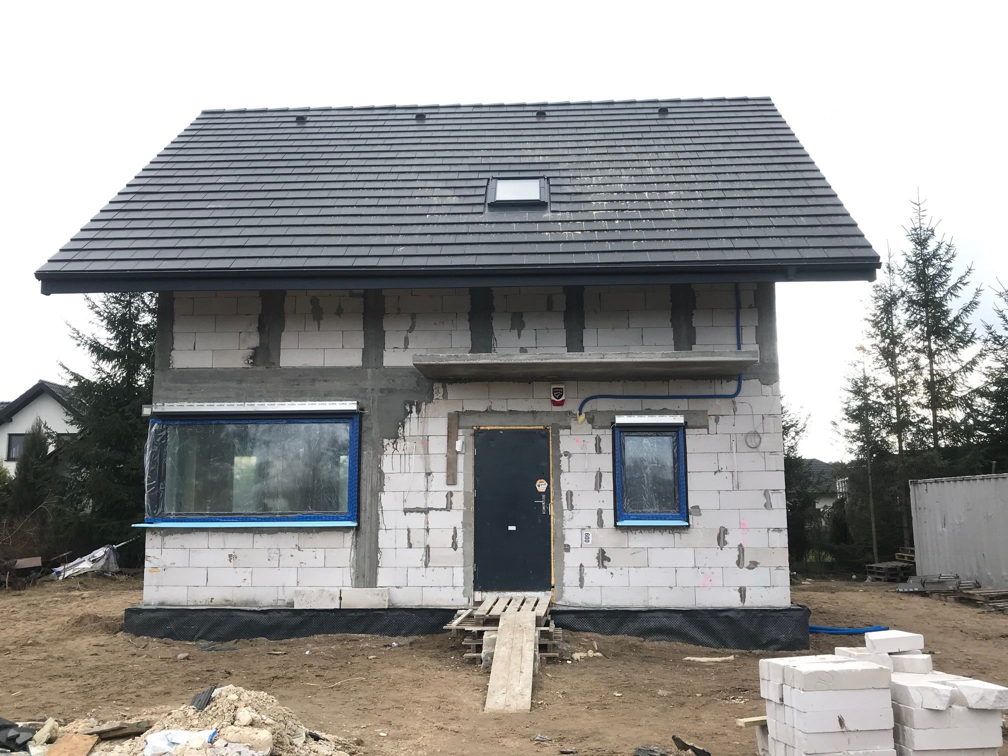 Dom w stanie surowym zamkniętym z ciemnoszarą dachówką ceramiczną, widoczne okna zabezpieczone folią, tymczasowe wejście z desek, białe bloczki konstrukcyjne.
