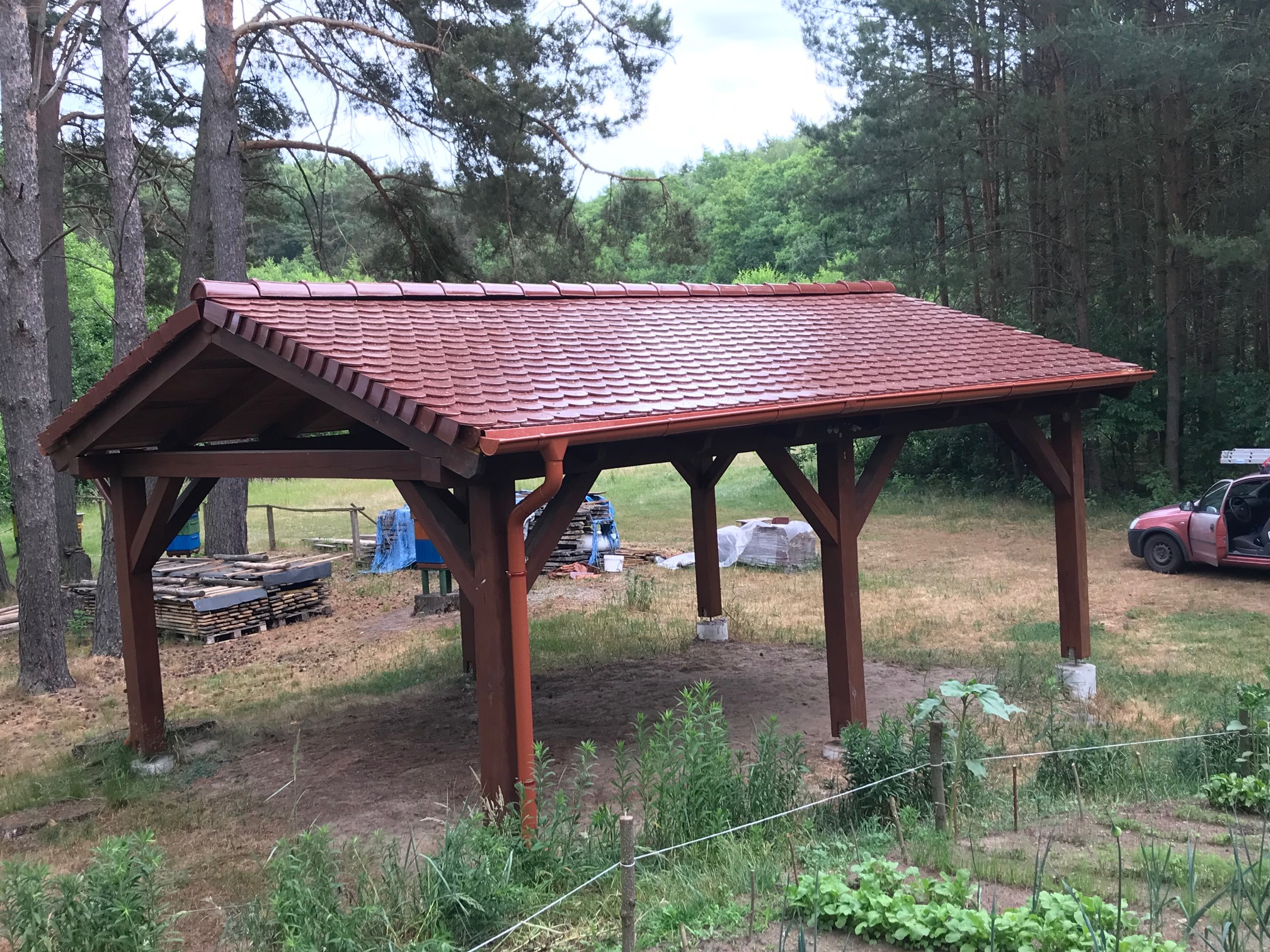 Drewniana altana z nowym, czerwono-brązowym dachem z dachówki ceramicznej, widoczna w otoczeniu drzew, z zapasem materiałów budowlanych w tle i zaparkowanym samochodem z otwartymi drzwiami.