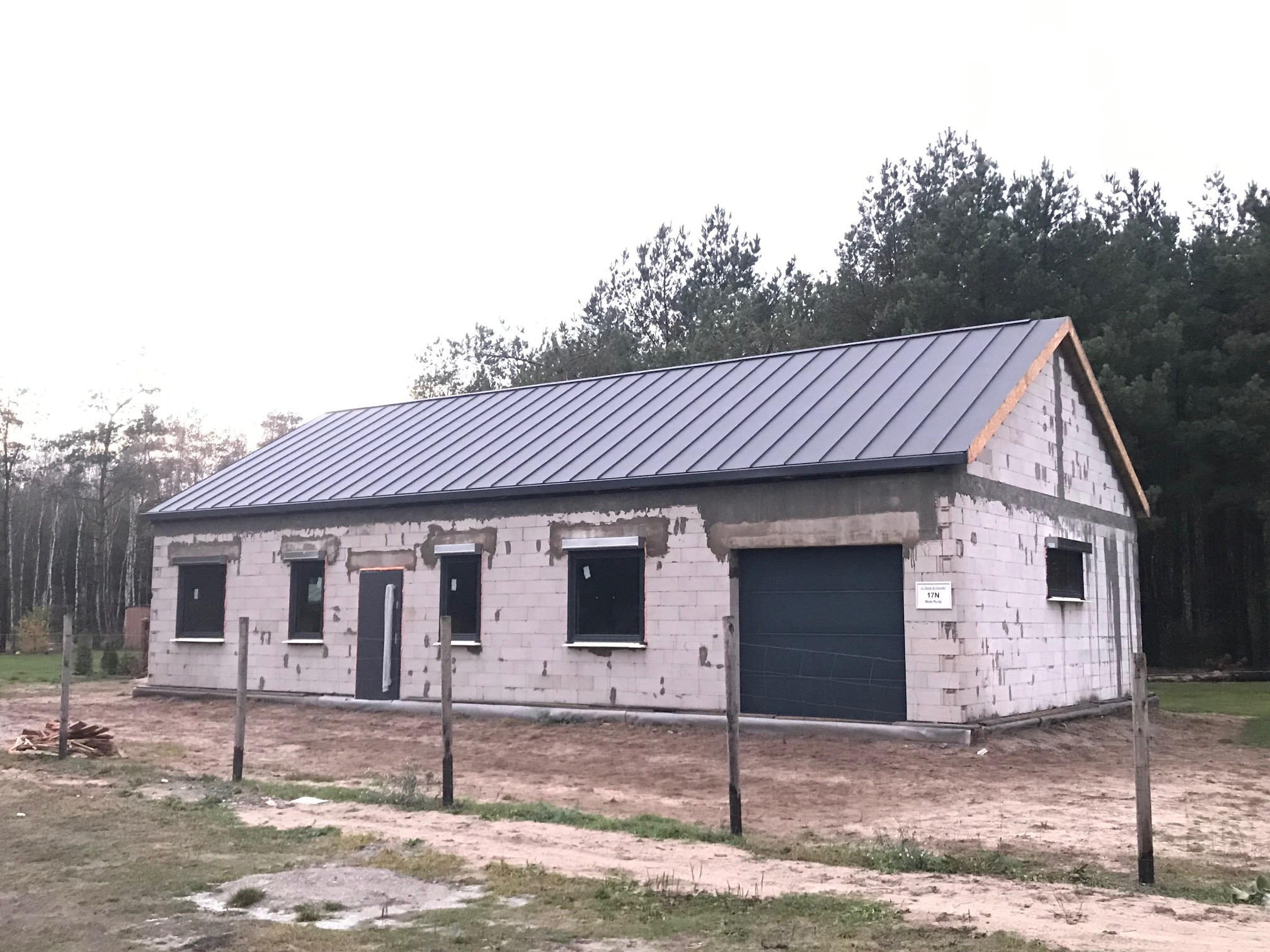 Surowy stan domu z szarego bloczka z zamontowanym ciemnoszarym dachem z blachy trapezowej i ciemnymi oknami, widok z zewnątrz na tle lasu, ogrodzenie z drewnianych pali.