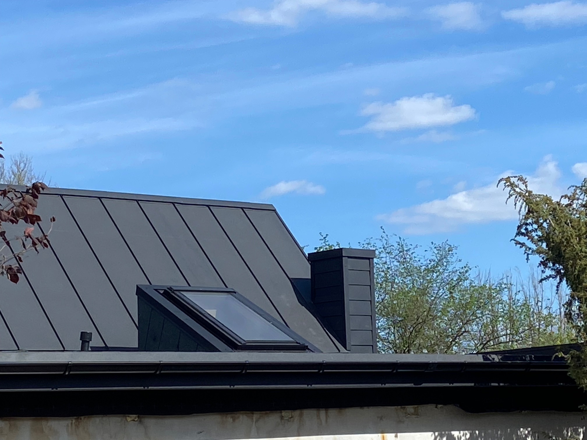 Ciemnoszary dach z blachy trapezowej z oknem dachowym i kominem na tle błękitnego nieba z chmurami.