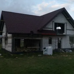 Budowa domu jednorodzinnego w trakcie, widoczny szkielet konstrukcji i częściowo ułożone brązowe blachodachówki na dachu, na pierwszym planie worki z materiałem budowlanym.