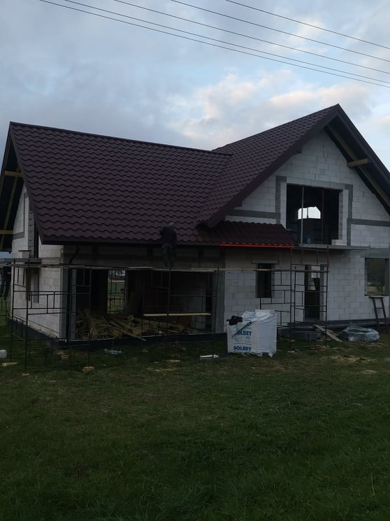 Budowa domu jednorodzinnego w trakcie, widoczny szkielet konstrukcji i częściowo ułożone brązowe blachodachówki na dachu, na pierwszym planie worki z materiałem budowlanym.