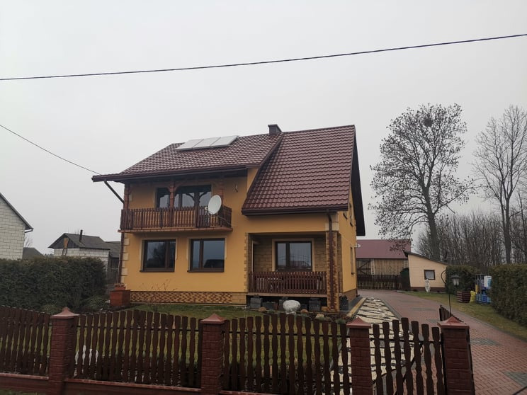 Dwupiętrowy dom z brązowym dachem z dachówki, balkonami z drewnianą balustradą i panelami słonecznymi na dachu, widziany zza drewnianego ogrodzenia.