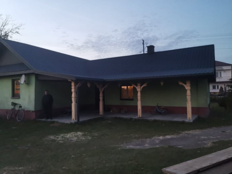 Zielony dom z ciemnym dachem z blachy trapezowej, drewnianą konstrukcją podtrzymującą okap i rowerem opartym o ścianę, widoczne obróbki blacharskie.