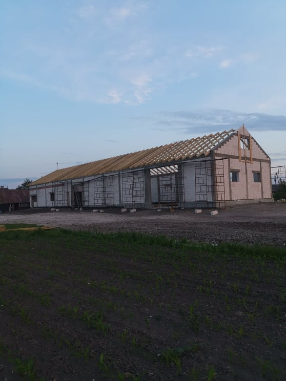 Budynek gospodarczy w trakcie budowy z częściowo ukończoną konstrukcją dachu, widoczne rusztowania i świeżo zasiana ziemia.