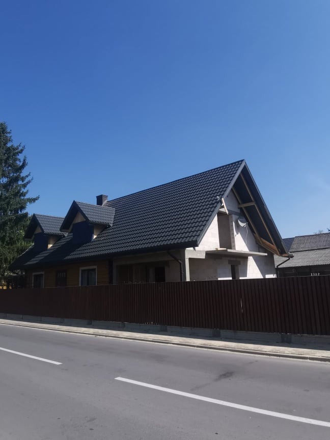 Dom jednorodzinny z ciemnoszarą, metalową dachówką, częściowo w trakcie budowy, widoczny zza brązowego ogrodzenia od strony ulicy w słoneczny dzień.