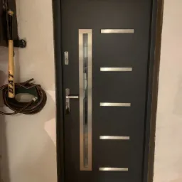 Ciemnoszare drzwi wejściowe z metalowymi wstawkami, zamontowane w surowym otoczeniu z widocznymi śladami prac wykończeniowych na ścianie i podłodze, obok ściany wisi kij bejsbolowy i zwinięty wąż...