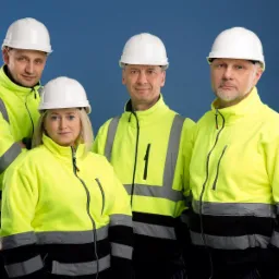 Grupa czterech osób, w tym jedna kobieta, ubranych w żółte bluzy robocze z elementami odblaskowymi i białe kaski ochronne, pozuje na jednolitym niebieskim tle.
