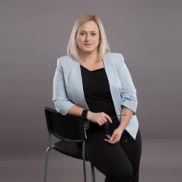 Portret kobiety w błękitnej marynarce opierającej się o czarne krzesło, trzymającej okulary w dłoni, na jednolitym szarym tle.