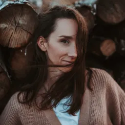 Portret kobiety w swetrze na tle stosu drewnianych bali, z włosami delikatnie przesłaniającymi twarz i subtelnym uśmiechem.