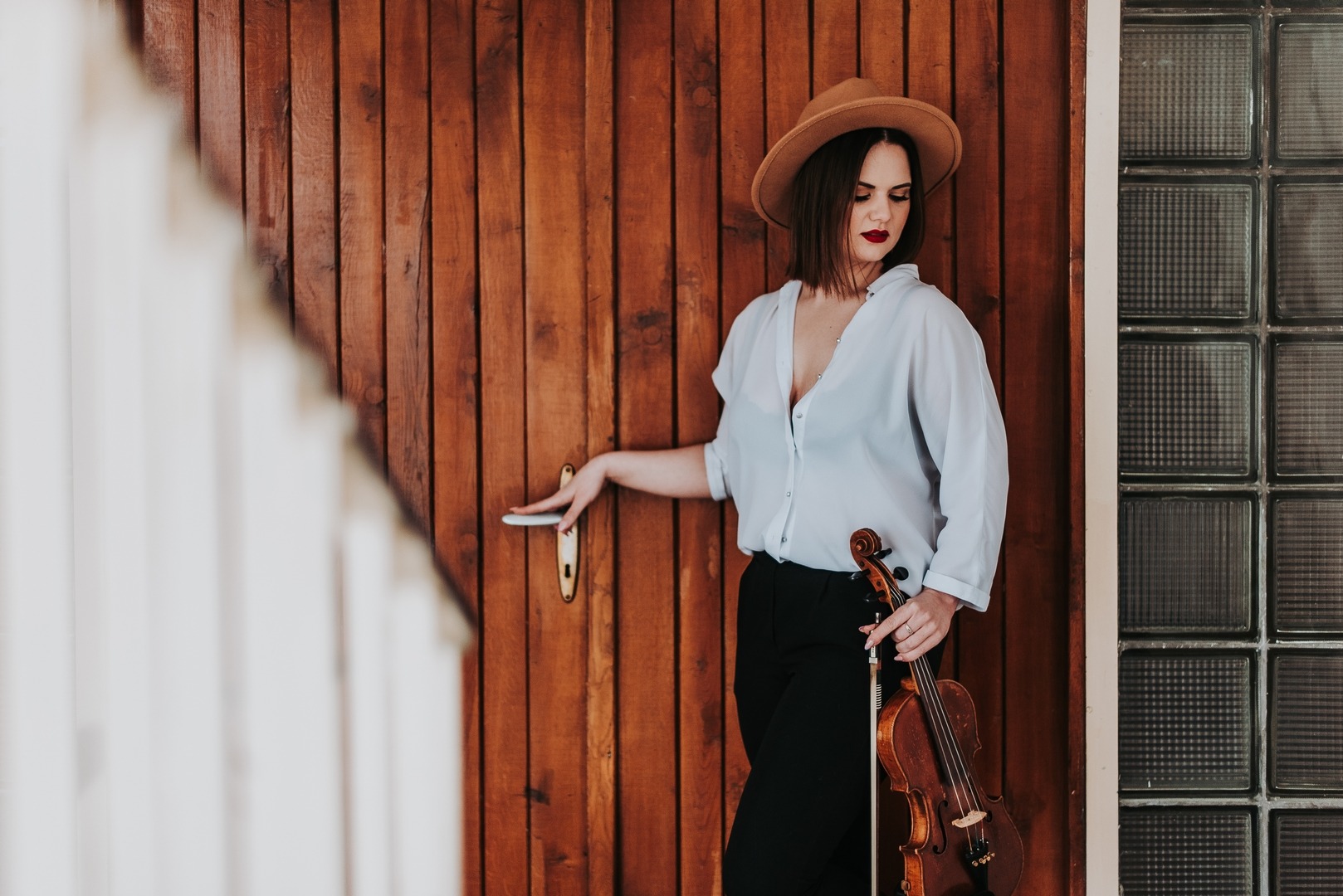 Kobieta w kapeluszu pozuje z skrzypcami przy drewnianych drzwiach i szklanych luksferach, ciemna szminka, biała koszula