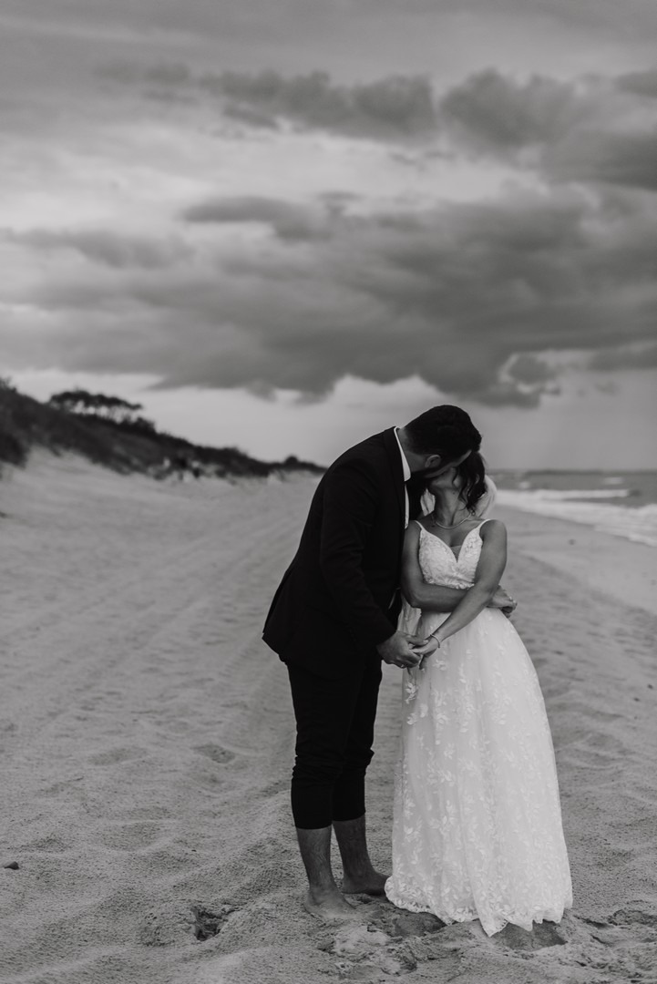 Czarno-białe zdjęcie pary młodej na plaży podczas pocałunku, z widocznym brzegiem morza i pochmurnym niebem w tle.