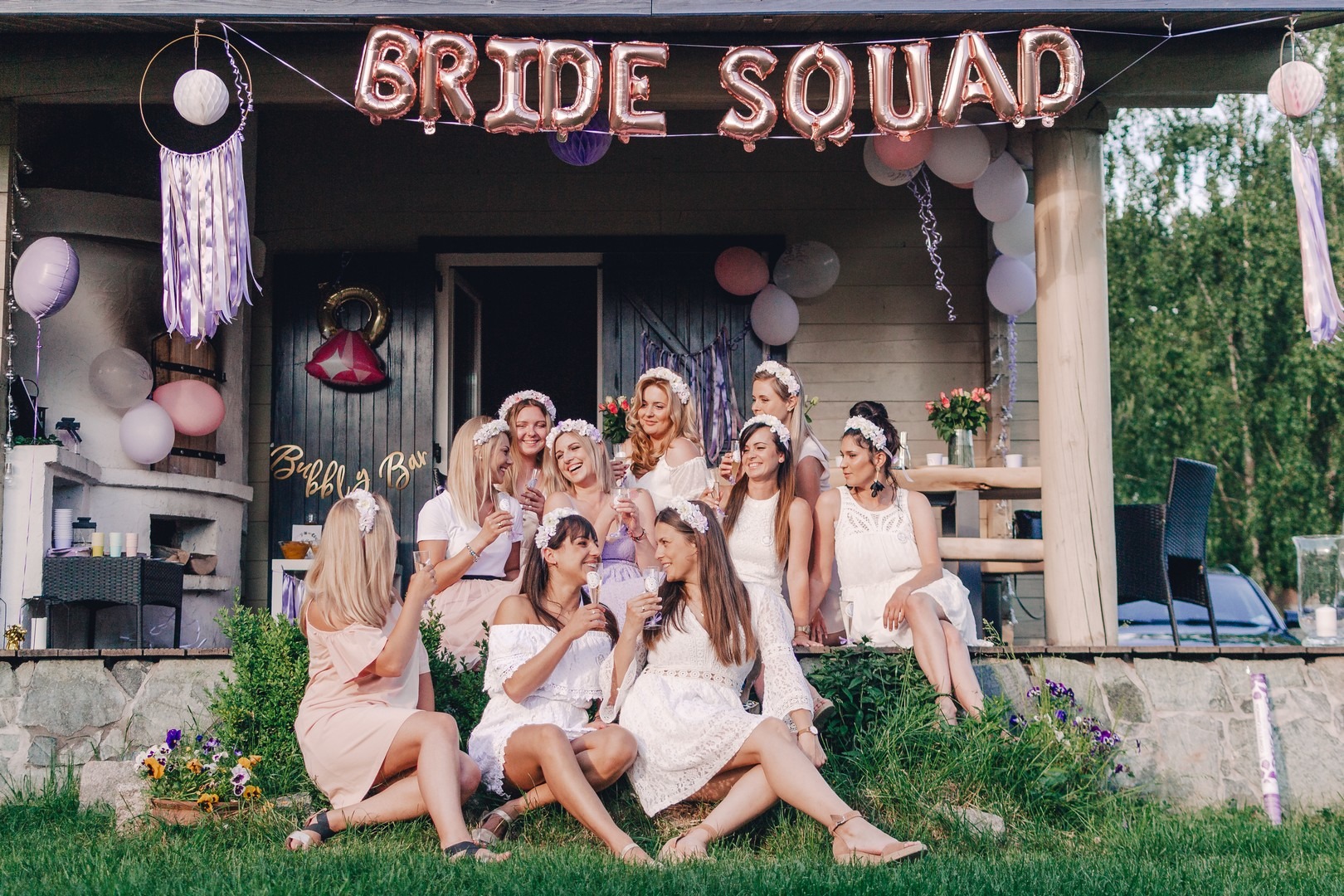 Grupa kobiet w białych sukienkach i kwiatowych wiankach pije szampana na trawie przed domem ozdobionym balonami 'BRIDE SQUAD', dekoracjami w odcieniach fioletu i napisem 'Bubbly Bar'.