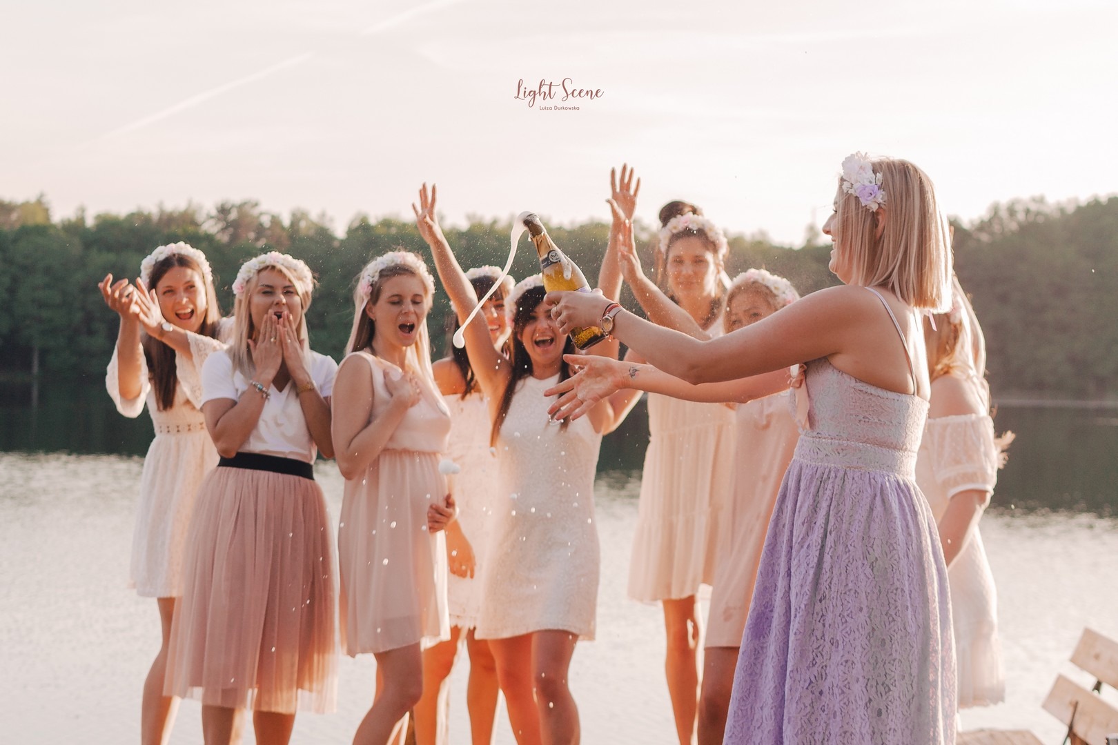 Grupa kobiet w jasnych sukienkach i kwiatowych wiankach świętuje, otwierając butelkę szampana nad jeziorem, z widocznym logotypem fotografa w górnym rogu.