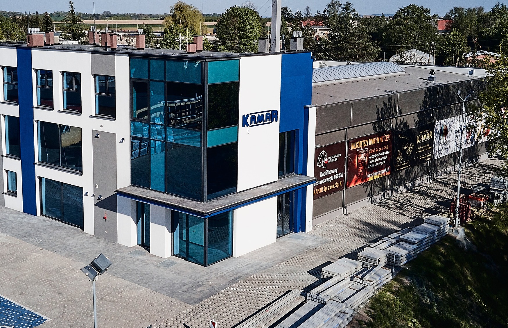 Nowoczesny budynek z logo Kamar, widoczne materiały budowlane na zewnątrz, plakaty reklamowe na ścianie bocznej.