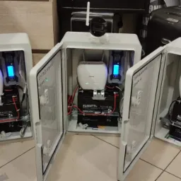 Trzy otwarte, białe szafki z zainstalowanymi kamerami, routerami i akumulatorami ALARMTEC, widoczne okablowanie, tło rozmyte.