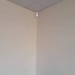 Biały czujnik alarmowy zamontowany w rogu pomieszczenia na tle jasnych ścian i sufitu kasetonowego.