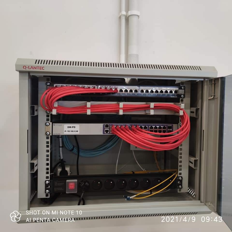 Otwarta szafa rack z zamontowanym switchem sieciowym Q-LANTEC, patch panelem i listwą zasilającą. Czerwone kable Ethernet uporządkowane za pomocą opasek zaciskowych. Niebieskie kable widoczne w tle.