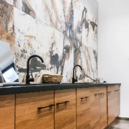 Łazienka z drewnianą szafką pod umywalką, czarnym blatem i armaturą, oraz ścianą wyłożoną płytkami imitującymi marmur. Na blacie koszyk z ręcznikami i małe radio.