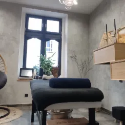 Nowoczesny gabinet masażu z wiszącym fotelem kokon, łóżkiem do masażu, drewnianymi szafkami i ścianą z mozaiki w odcieniach szarości.