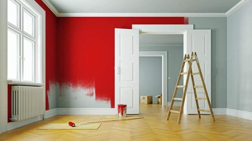 Świeżo pomalowana na czerwono ściana w pokoju, widoczna drabina oparta o białe drzwi, puszka z farbą i wałek malarski leżące na podłodze w jodełkę, fragment ściany w szarym kolorze.
