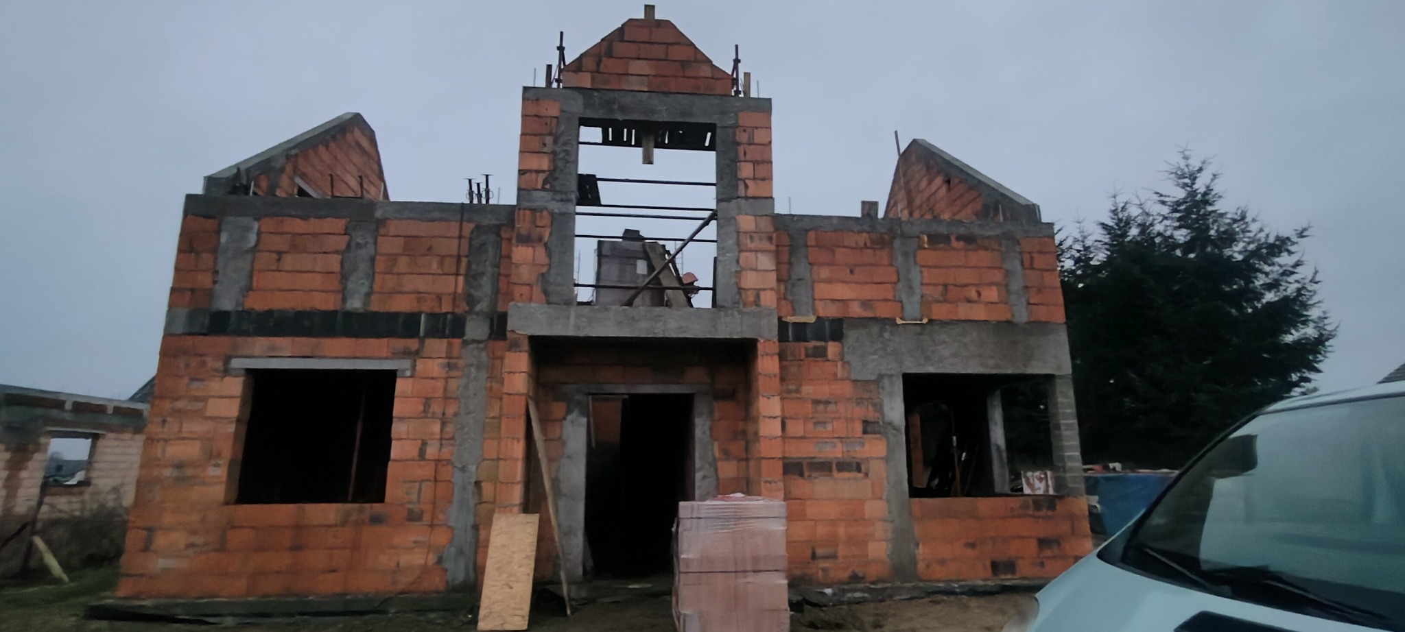 Surowy stan domu z czerwonej cegły z widocznymi otworami okiennymi i drzwiowymi, częściowo zabetonowanymi nadprożami. W tle widoczny fragment lasu i samochód.