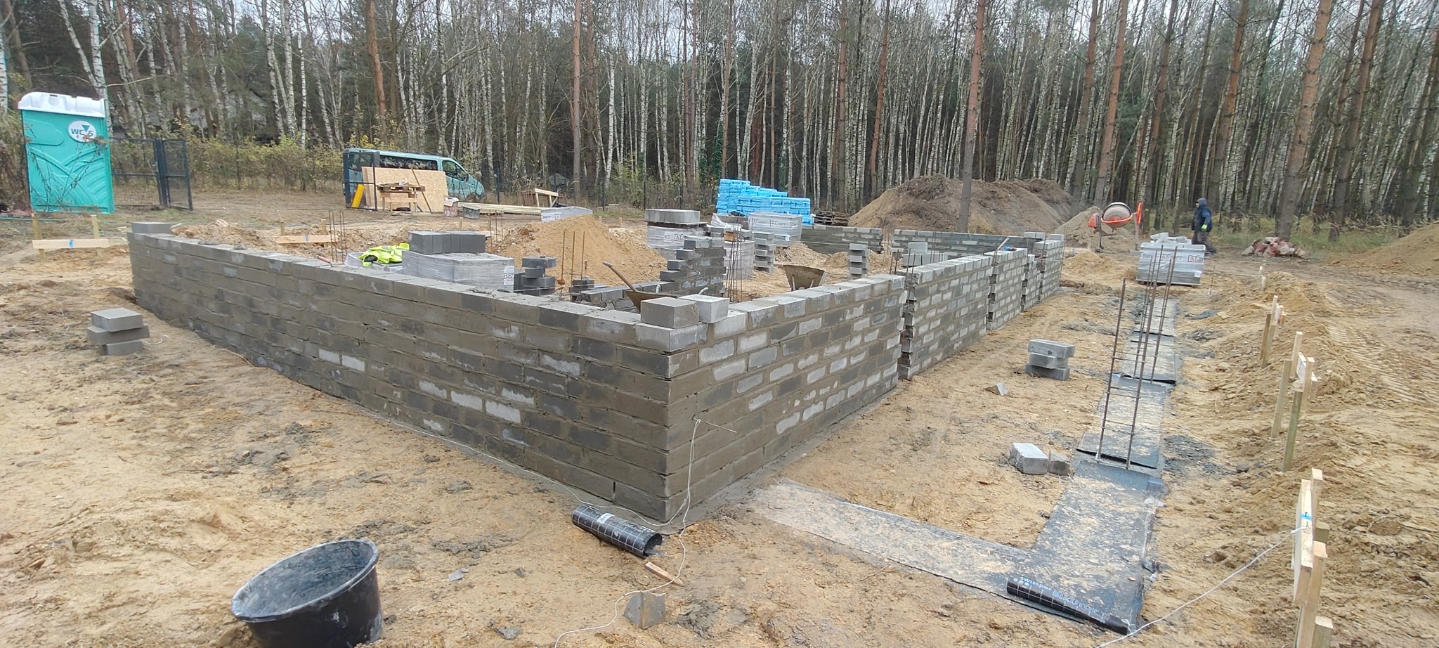 Wznoszenie murów fundamentowych z bloczków betonowych na piaszczystym terenie budowy. Widoczne zbrojenia i przygotowane materiały. W tle las i zaplecze budowy.