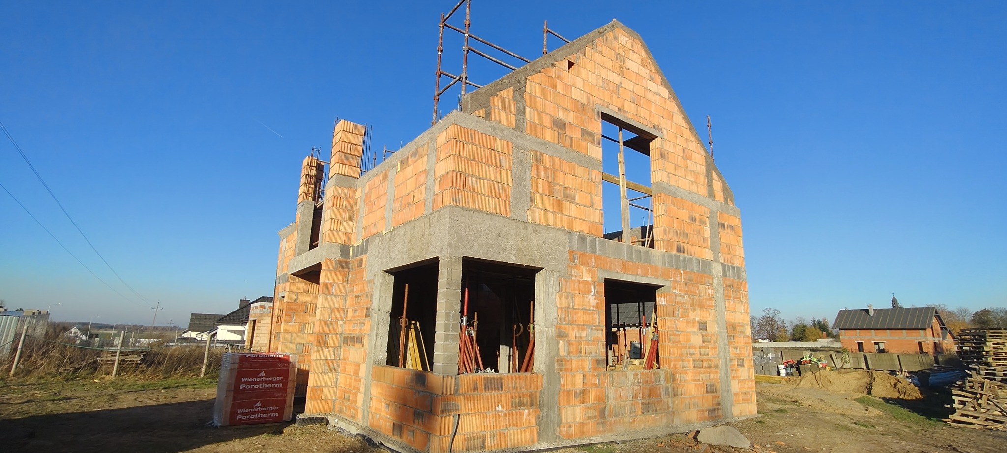 Wznoszenie ścian domu z czerwonej cegły Porotherm, widoczne otwory okienne i zbrojenia, na pierwszym planie paleta z cegłami, błękitne niebo w tle.