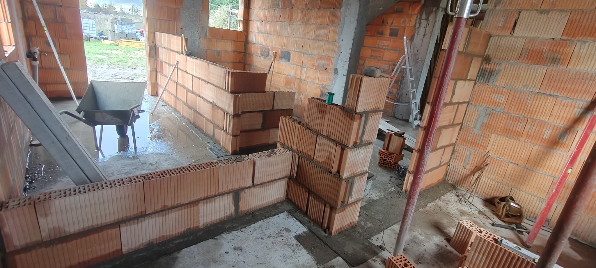 Wnętrze w trakcie murowania ścian z czerwonej cegły dziurawki. Widoczne narzędzia, taczka i poziomica. Częściowo wymurowane ściany działowe i nośne.