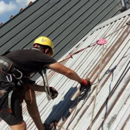 Pracownik w kasku i uprzęży zabezpieczającej na pochyłym, metalowym dachu w Krakowie, zabezpieczony linami, wykonuje prace na wysokości.