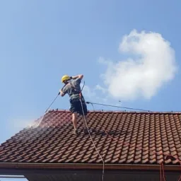 Pracownik w uprzęży i kasku myje dach z czerwonej dachówki za pomocą myjki ciśnieniowej; widoczna piana i strumień wody pod ciśnieniem.