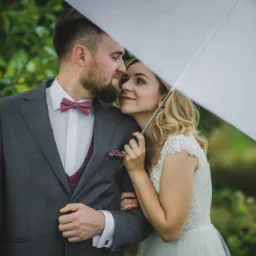 Para młoda pod białym parasolem: mężczyzna w szarym garniturze z bordową muchą i kamizelką, kobieta w koronkowej sukni, oboje patrzą na siebie z czułością.