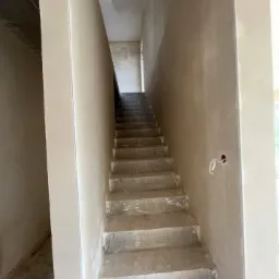 Surowe schody betonowe prowadzące w górę w budynku w trakcie wykańczania, ściany częściowo pokryte jasną warstwą wykończeniową, widoczne niedoskonałości i przewody.