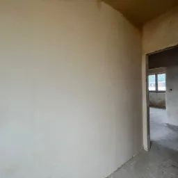 Świeżo nałożona, jasna gładź na ścianie w rogu pomieszczenia, widoczny surowy betonowy sufit i przygotowane do wykończenia przejście do innego pokoju z oknem.